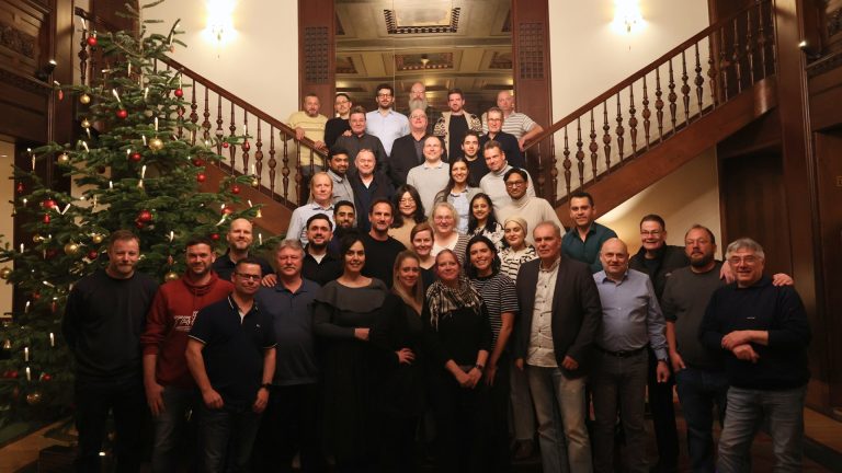 Mitarbeitende der redi-Group stehen gemeinsam vor einem geschmückten Weihnachtsbaum in einem historischen Treppenhaus.